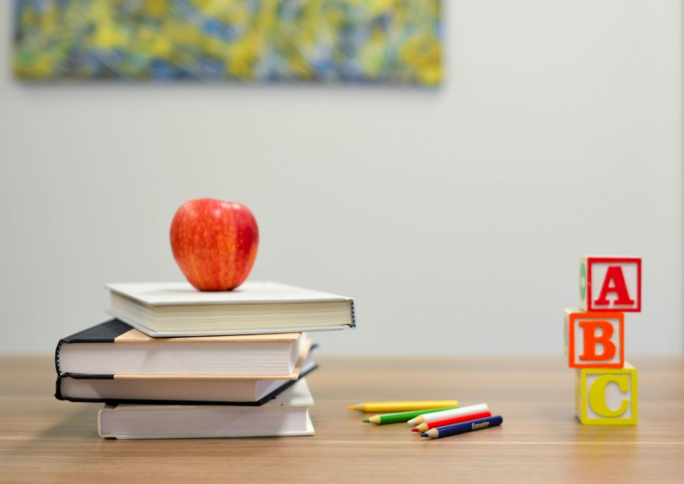 A stack of books with an apple on top, some colored pencils, and blocks reading ABC on a table